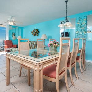 Bright turquoise walls complement a dining area with a glass-top table, surrounded by six chairs and views of the beach