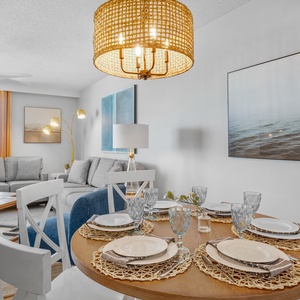 Bright dining area featuring a round table set for four, with a cozy living space and beach view through large windows
