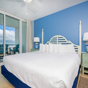 The master bedroom features a comfortable bed, blue accent wall, and a stunning beachfront view from the sliding glass doors