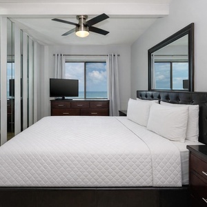 The master bedroom features a plush king bed, a large window with a stunning beachfront view, and modern furnishings