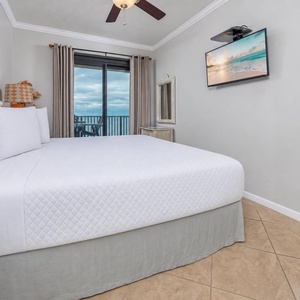 Master suite bedroom featuring a comfortable king bed, a mounted TV, and access to a balcony with a view of the beach