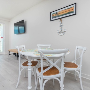 This dining area features a round table with white chairs, a wall-mounted TV, and a stunning beachfront view