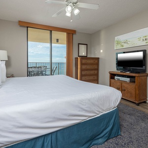 Master bedroom featuring a large king bed and a TV on a wooden stand, leading to a balcony with a beach view