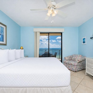 Bright blue walls complement a spacious room with a king bed, cozy floral chair, and balcony access showcasing a beach view