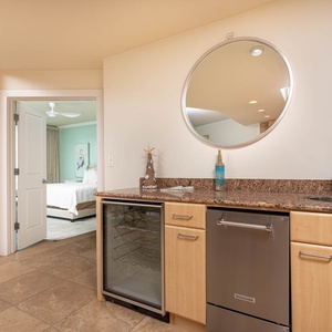 This kitchen features a sleek wet bar with a wine cooler, stylish granite countertops, and a view into the inviting bedroom