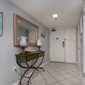 Welcoming entryway features a green console table with decorative lamps, a large mirror, and coastal-themed wall art