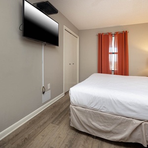 A cozy guest bedroom features a king bed with white linens, twin bunk bed, an orange curtain, and a wall-mounted TV