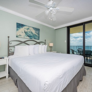 A king bed with white linens is complemented by coastal artwork and leads to the balcony with a view of the beach front