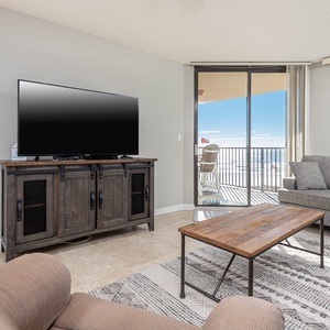 The living room features a cozy gray sofa, a rustic coffee table, and a large TV, with expansive views of the beach beyond
