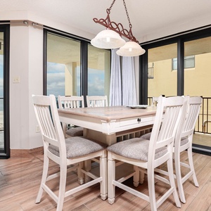 The dining area features a wooden table surrounded by cushioned chairs, with large windows showcasing a beach front view