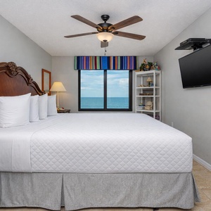 The master bedroom features a plush king bed, a mounted TV, and a stunning beachfront view from the large window