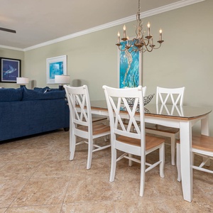 Dining area featuring a glass-top table with white chairs, a blue sofa nearby, and artwork enhancing the coastal vibe