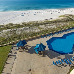 The view showcases the sparkling pool area with blue lounge chairs and shaded cabanas, overlooking a pristine beach