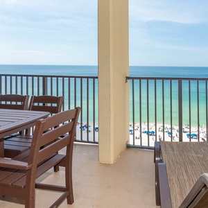 A spacious balcony features a patio set and lounge chair, offering a direct view of the beach and turquoise waters