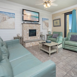 Living area features a cozy teal sofa, two matching chairs, and a fireplace, with a view of the beach through large windows