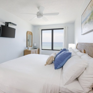 This bedroom features a bed with decorative pillows, a wall-mounted TV, and stunning beachfront views through large windows