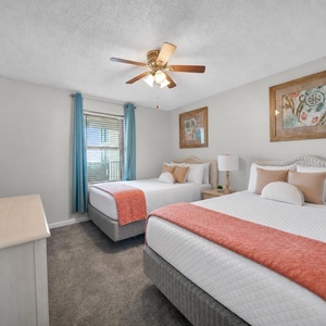 Bright guest bedroom featuring two double beds with coral throws, a TV, and large windows framed by blue curtains