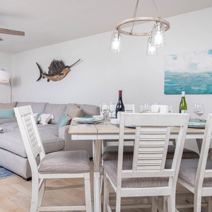 Cozy dining area features a light wood table set for six, with a view of the beach visible through sliding glass doors
