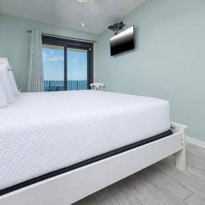 Bright master bedroom featuring a comfortable king bed, a mounted TV, and a balcony door revealing a serene view of the beach