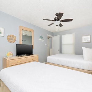 A guest room features two beds, a TV on a wooden dresser, and light blue walls adorned with beach decor