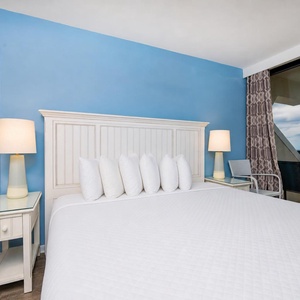 This master bedroom features a plush king bed with white linens, a light blue accent wall, and a balcony view of the beach