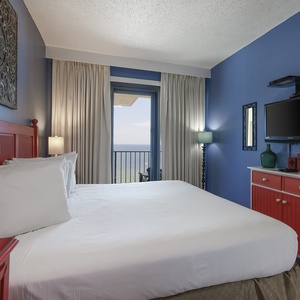 This vibrant bedroom features a red accent wall, a cozy bed, a mounted TV, and a stunning view from the balcony