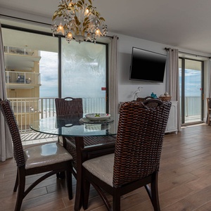 This dining area features a glass table and woven chairs, with a view of the beach and a wall-mounted TV for entertainment