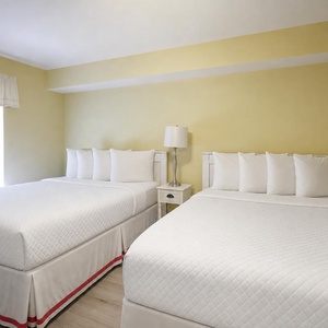 Guest room featuring two queen beds with crisp white linens, a bedside lamp, and natural light from the window