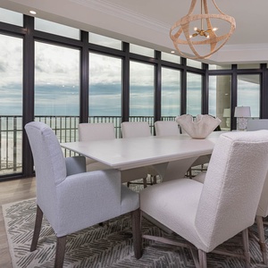 The dining area features a large table with ten soft, light-colored chairs, with panoramic beach views through large windows