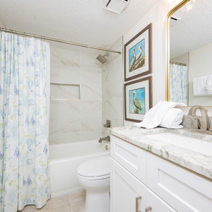 Trndy guest bathroom featuring a tub/shower combo, elegant granite countertops, and coastal-themed artwork on the walls