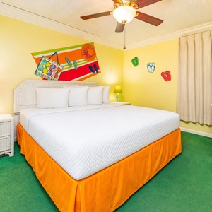 Brightly decorated bedroom featuring a large king bed with white linens, colorful accents, and a TV on a white dresser