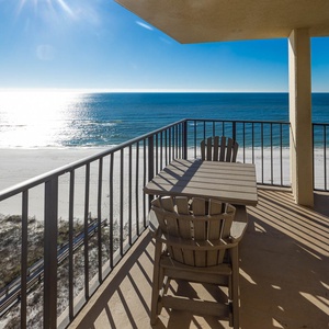A private balcony features a wooden table and chairs, offering a serene view of the shimmering water and white sand below