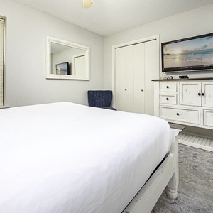 Elegant guest bedroom featuring a queen bed, a cozy blue chair, and a TV mounted above a white dresser