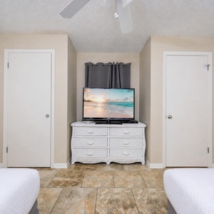 A cozy guest room features two double beds, a white dresser, and a TV displaying a serene beach scene