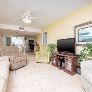 The living area features comfortable seating, a TV, and a glimpse of the dining space and kitchen beyond