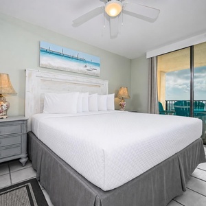 Brightly decorated master bedroom featuring a king bed, coastal artwork, and access to a balcony overlooking the beach