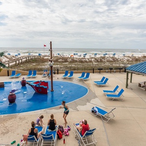 Vibrant pool area features a playful pirate ship splash park and lounge chairs, with a beach view of colorful umbrellas
