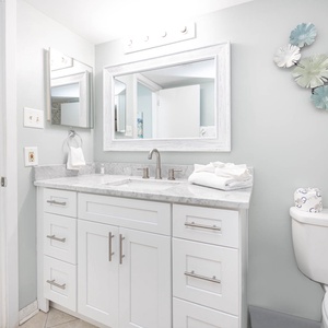 A bright bathroom features a white vanity with marbled countertop, decorative mirrors, and a cozy living area visible beyond