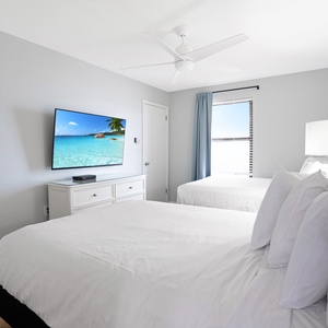 Guest bedroom features two full beds, a mounted TV, and a window with a view of the beach front and palm trees