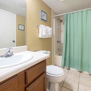 The bathroom features a clean design with a tub/shower combo, fresh towels, and a bright green shower curtain