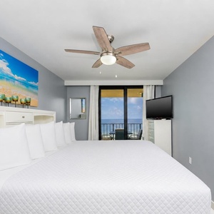Bright master bedroom featuring a king bed, TV, and sliding doors leading to the balcony with a beach front view