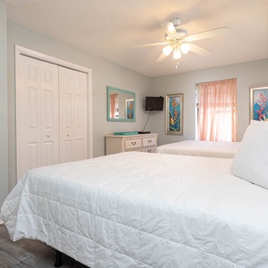 Bright guest bedroom featuring two double beds, a dresser, and colorful art, with a ceiling fan and natural light