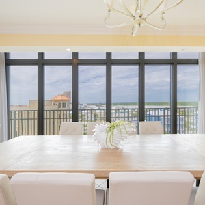 Bright dining area with a large wooden table, white chairs, and expansive windows showcasing a beautiful coastal view