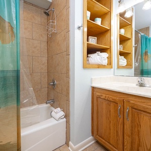 Refreshing guest bath features a tub/shower combo with a beach curtain, wooden cabinetry, and neatly arranged towels