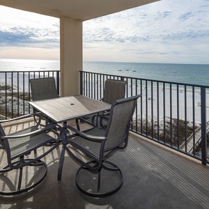 The balcony features a table and chairs, offering a serene view of the beach and gentle waves beyond the railing