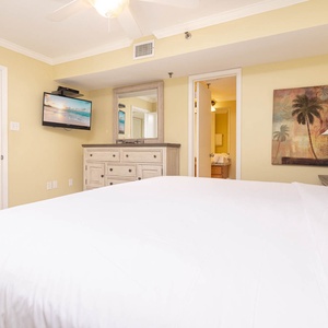 The cozy bedroom features a plush king bed, a mounted TV, and a dresser, with a warm yellow ambiance and artwork