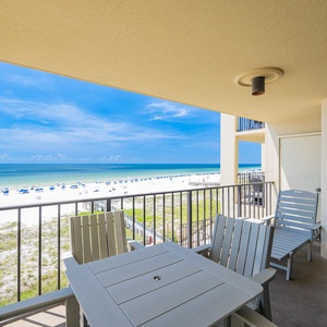 The balcony features a table and chairs, offering a clear view of the beach and vibrant blue waters beyond