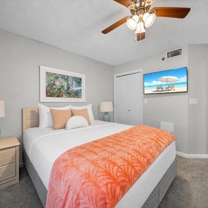 Brightly decorated guest bedroom featuring a queen bed with an orange throw, a bedside lamp, and a wall-mounted TV