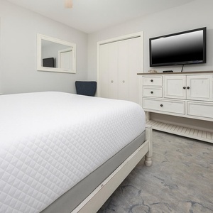 Elegant guest bedroom featuring a queen bed, a cozy blue chair, and a TV mounted above a white dresser