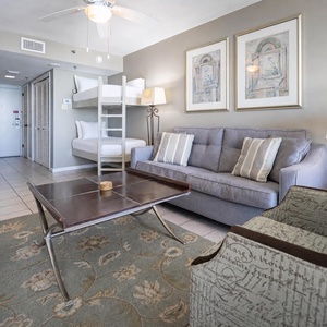 A cozy living area features a gray sofa with queen sleeper, decorative armchair, and twin bunk beds against a light wall